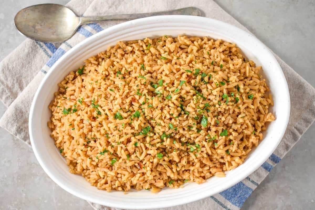 The seasoned rice served in a white, oval dish and set on a beige and blue linen with a serving spoon on the top.