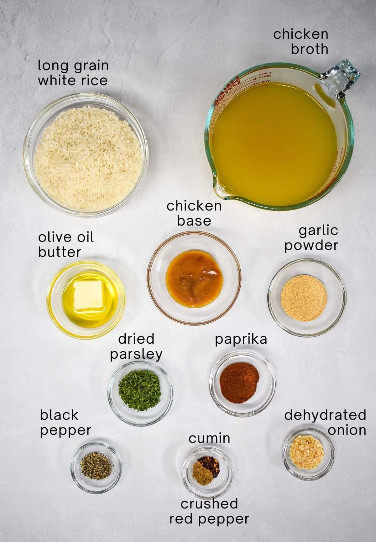 The ingredients for the rice arranged in glass bowls on a white table. Each ingredient is labeled in small, black letters.
