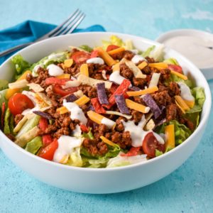 The ground beef taco salad served in a white bowl set on an aqua colored table with a blue linen, a form, and a small dish with dressing in the background.