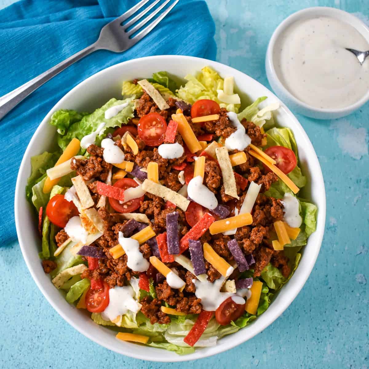 The ground beef taco salad served in a white bowl set on an aqua colored table with a blue linen, a form, and a small dish with dressing in the background.