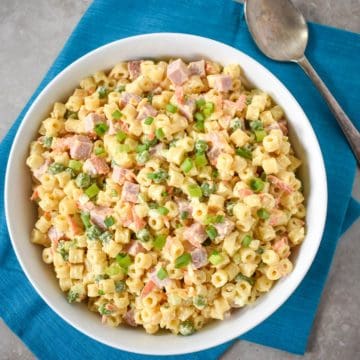 The finished ham pasta salad in a large, white bowl set on a teal linen with a serving spoon to the right.