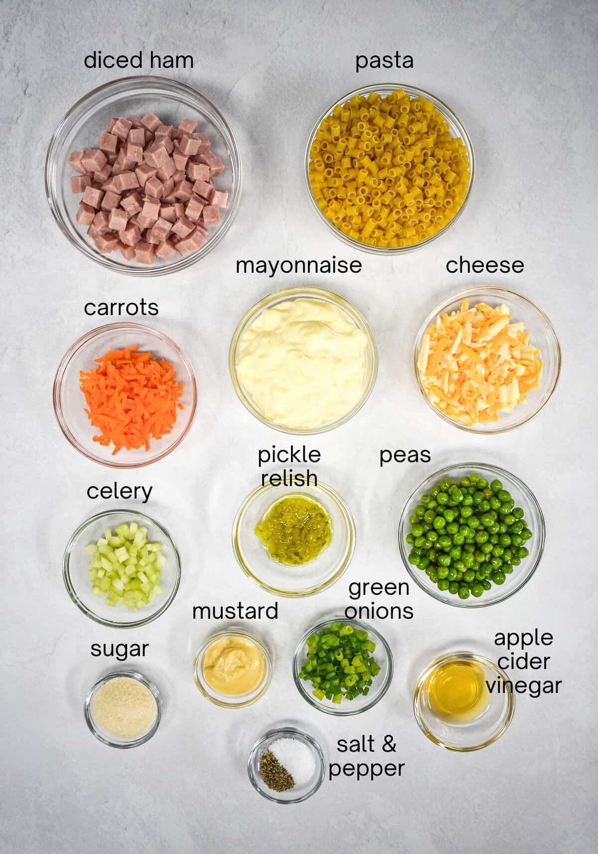 The ingredients for the ham pasta salad prepped and arranged in glass bowls on a table. Each one is labeled with small black letters.