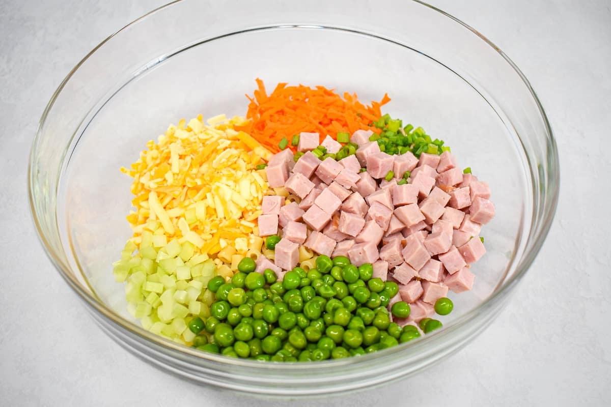 Cooked pasta covered by diced ham, shredded carrots, cheese, celery, peas, and green onions in a large, glass bowl.