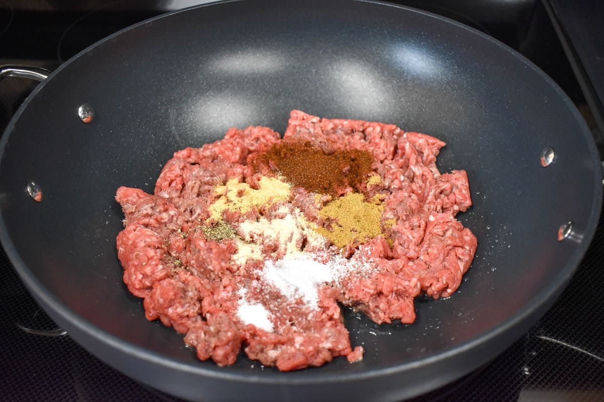 Raw ground beef with seasoning on top in a large, black skillet.