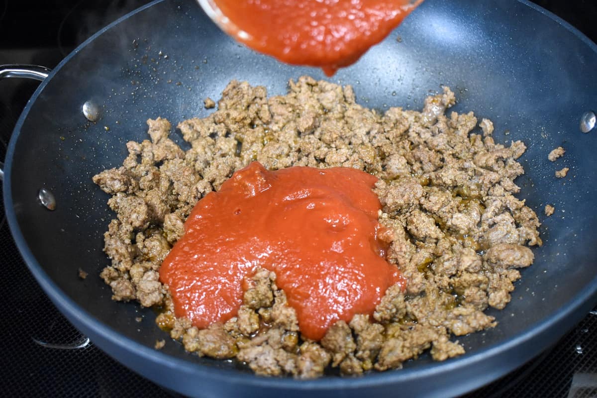 Tomato sauce being added to browned ground beef in a large, black skillet.