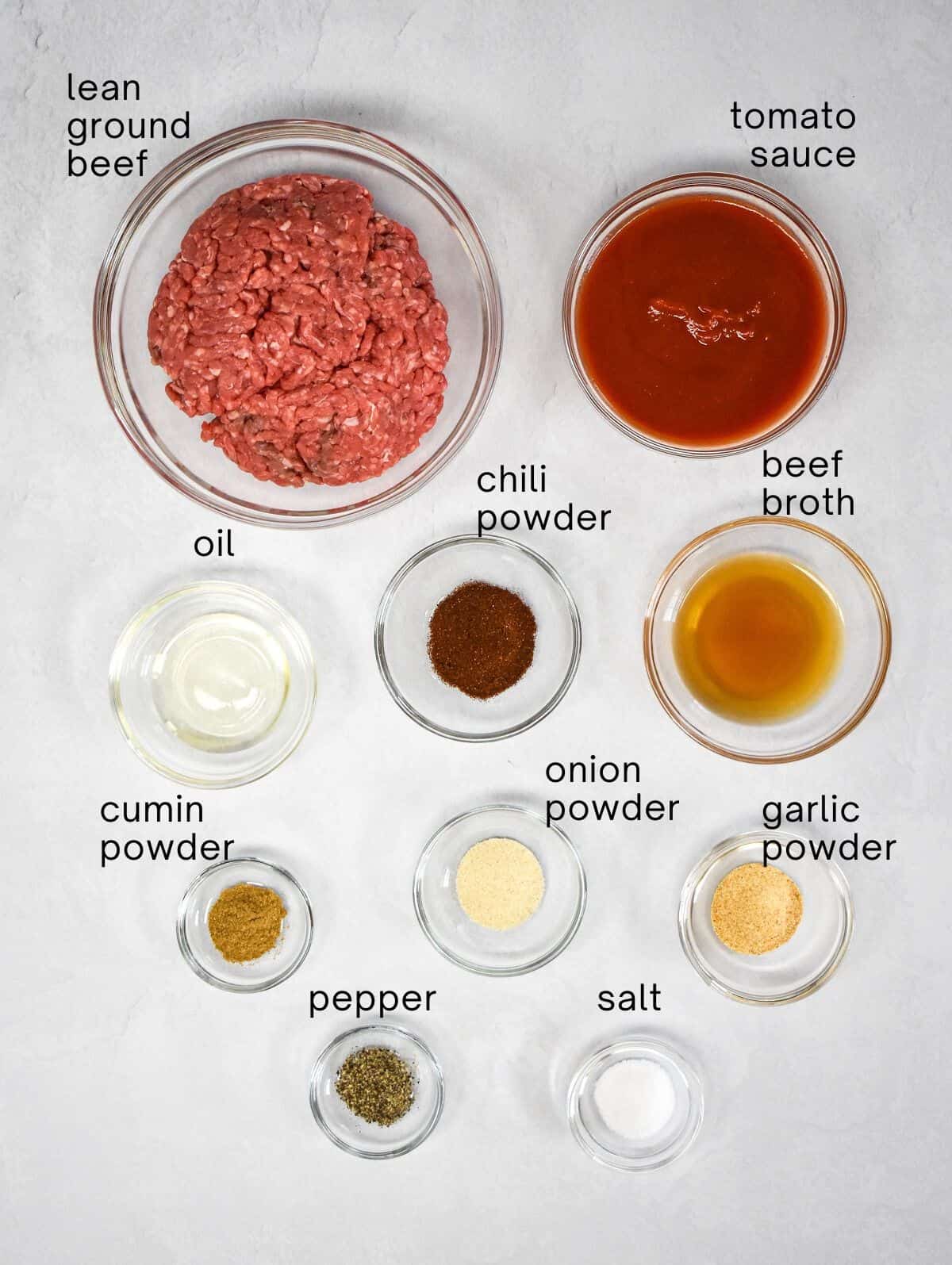 The ingredients for the taco meat for the salad arranged in glass bowls and set on a white table. Each ingredient is labeled in small black letters.