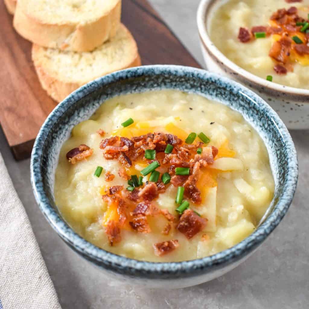 The potato soup topped with shredded cheese, bacon bits and chives with sliced bread and other soup in the background.