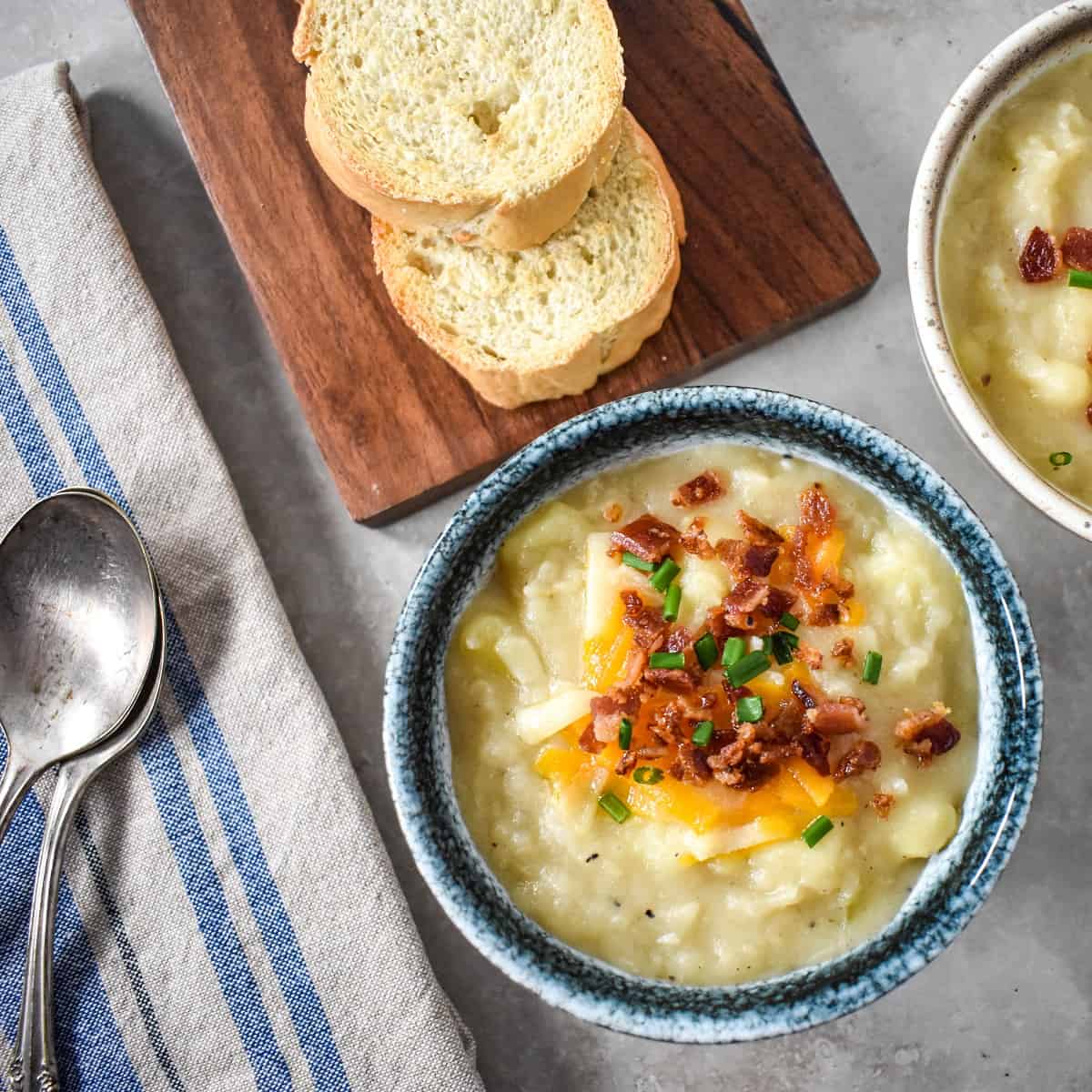 The potato soup topped with shredded cheese, bacon bits and chives with sliced bread and other soup in the background.