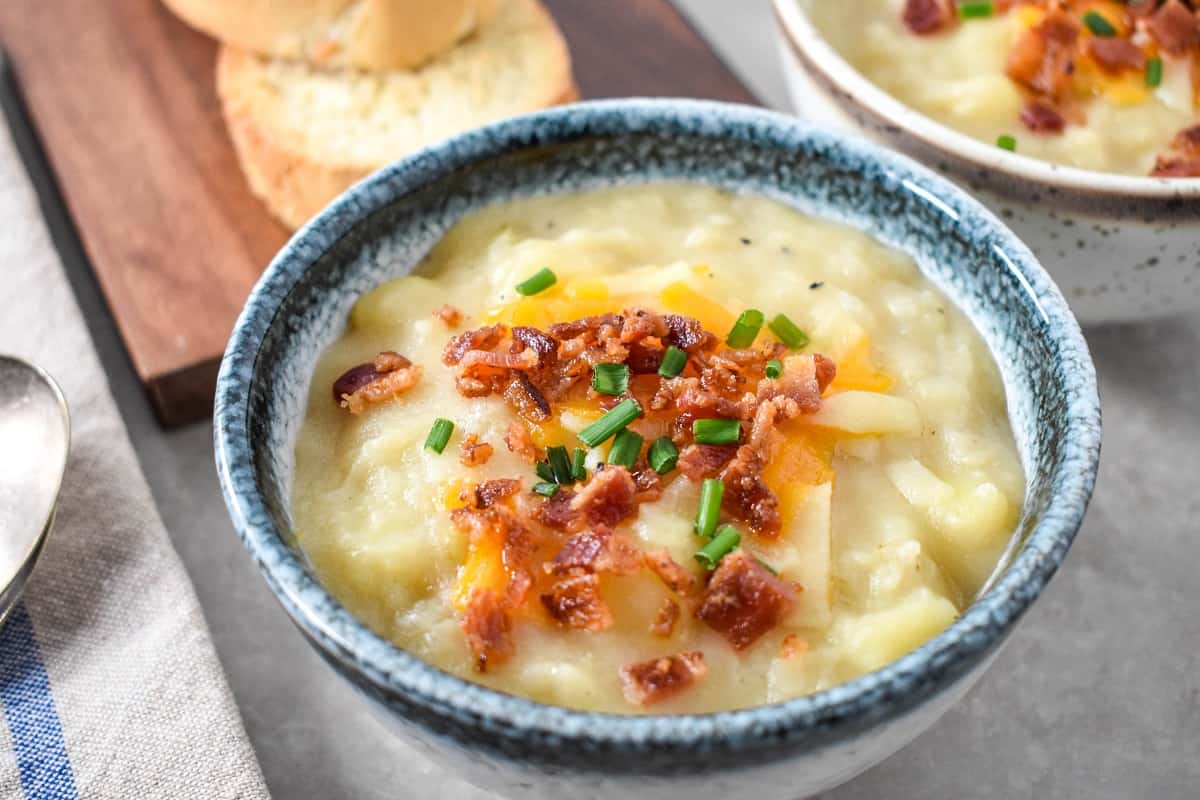 A close up of the finished potato soup served in a white bowl with a blue rim with bread slices and a another soup visible in the background.