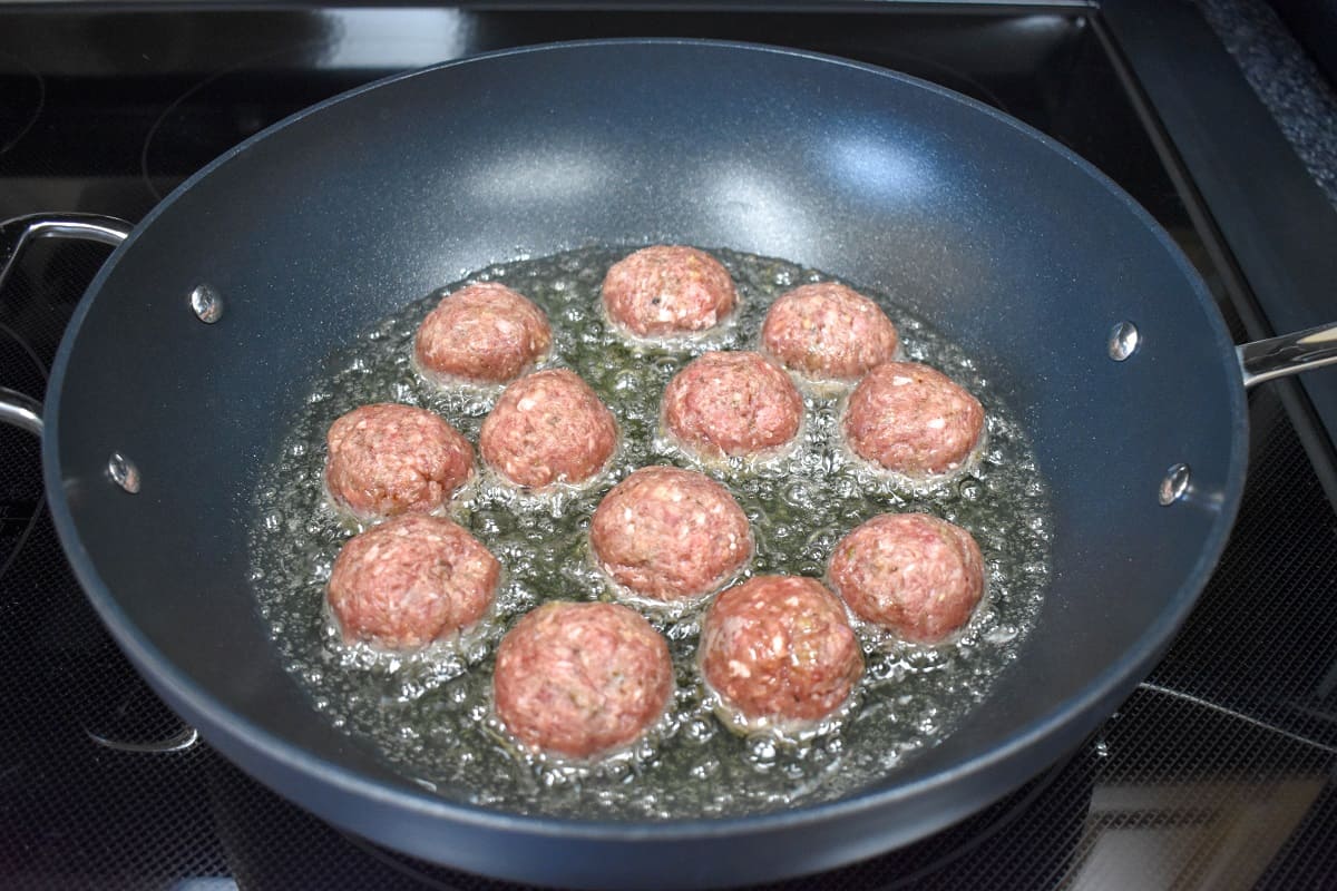 Meatballs cooking in oil in a large, non-stick skillet.