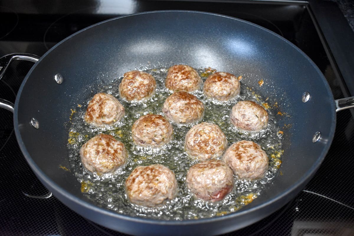 Meatball browned on one side cooking in oil in a large, non-stick skillet.