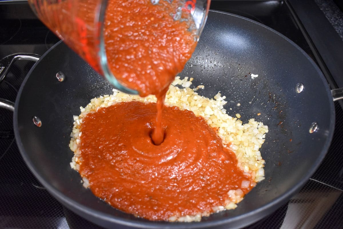 Pasta red sauce being added to the onions and garlic in a large skillet.