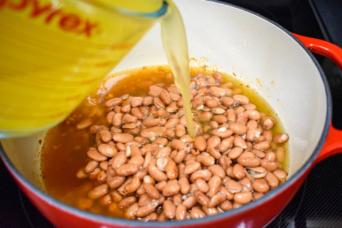 Chicken broth being added to pinto beans in a large, white and red pot.