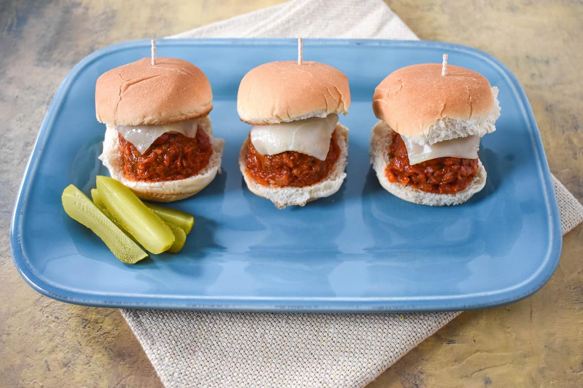 Three sliders served on a light blue plate with pickle wedges on the left side.