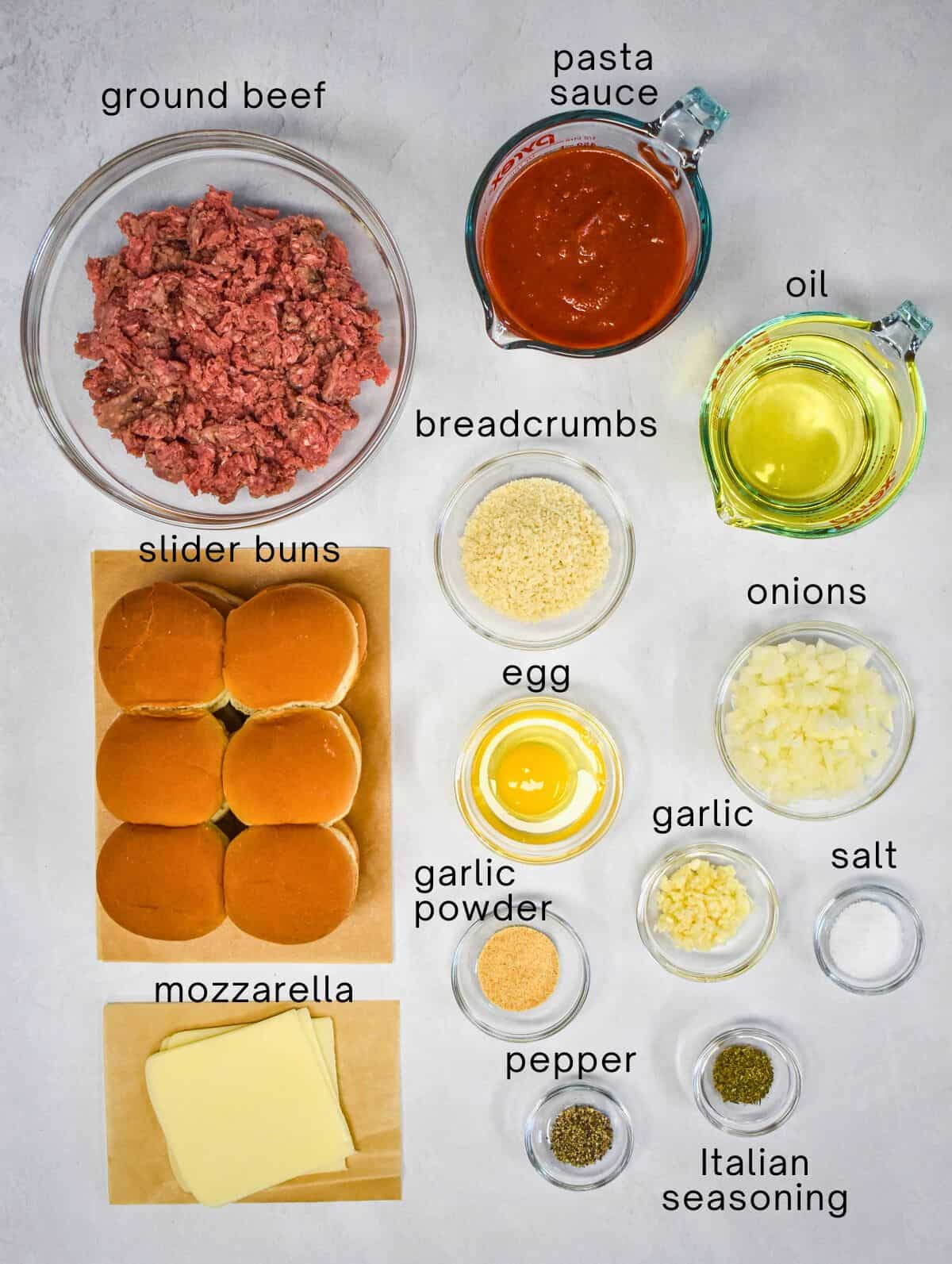 The ingredients for the sliders prepped and arranged on a white table with each labeled with small black letters.