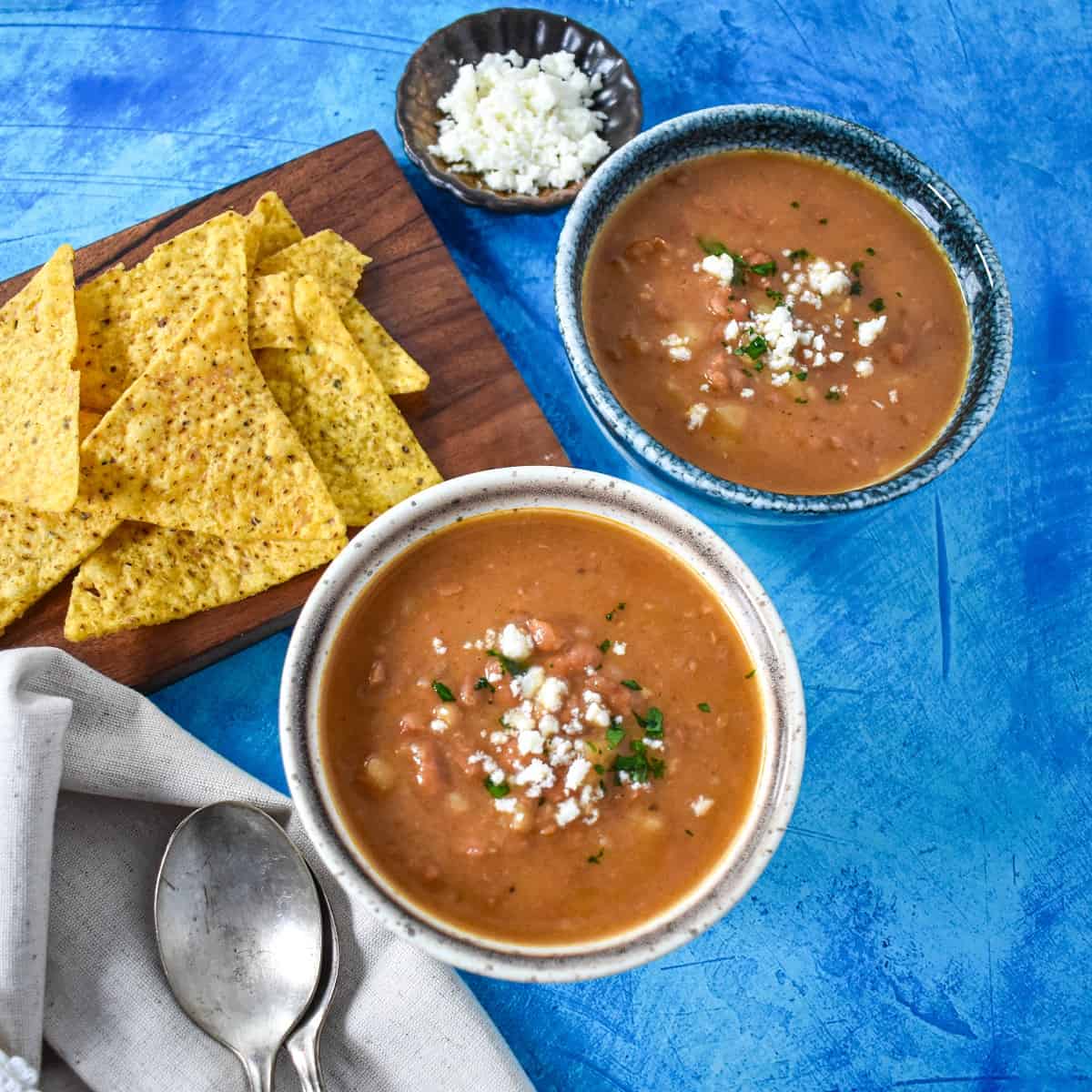Two bowls of pinto bean soup served with tortilla chips and queso fresco on the side all set on a blue table.