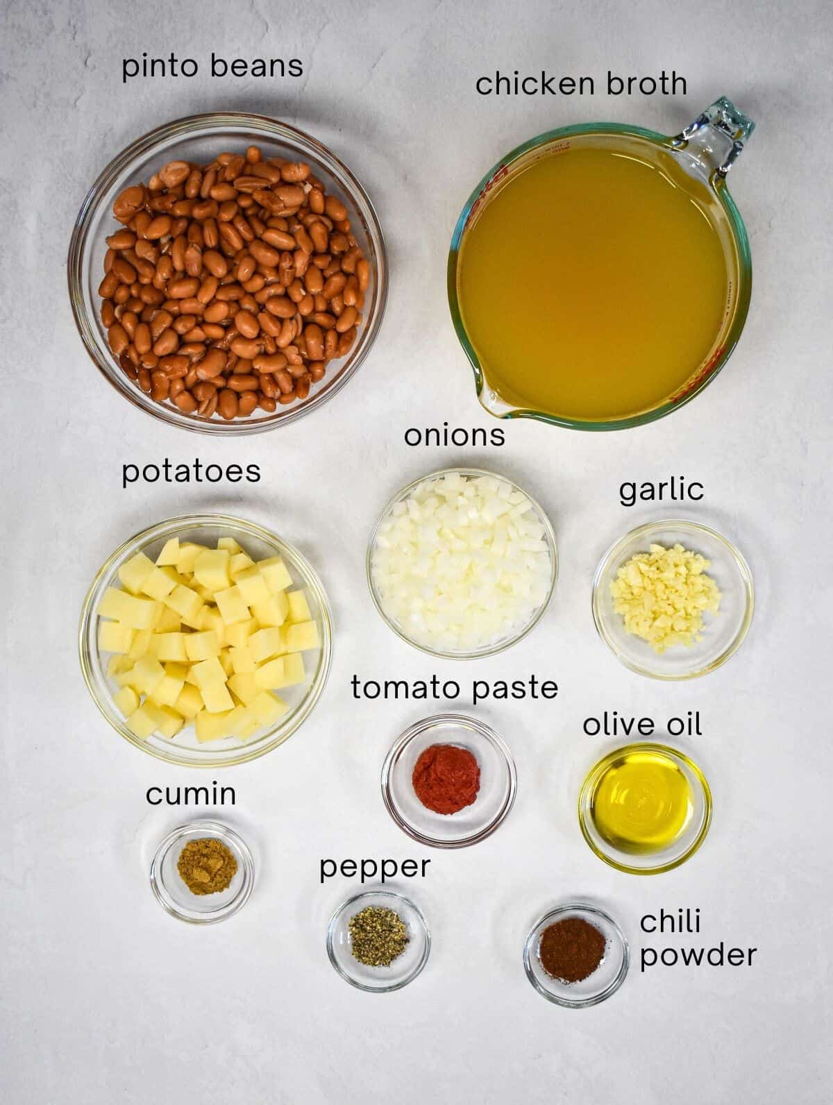 The ingredients for the soup prepped and arranged in glass bowls on a white table with each labeled with small, black letters.