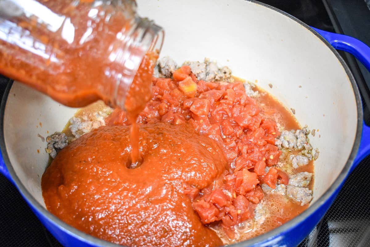 Pasta sauce being added to diced tomatoes and browned sausage in a large white and blue pot.