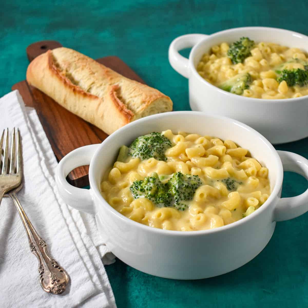 Two white bowls with the broccoli macaroni and cheese set on a green table with a piece of bread and white napkins and forks to the left side.