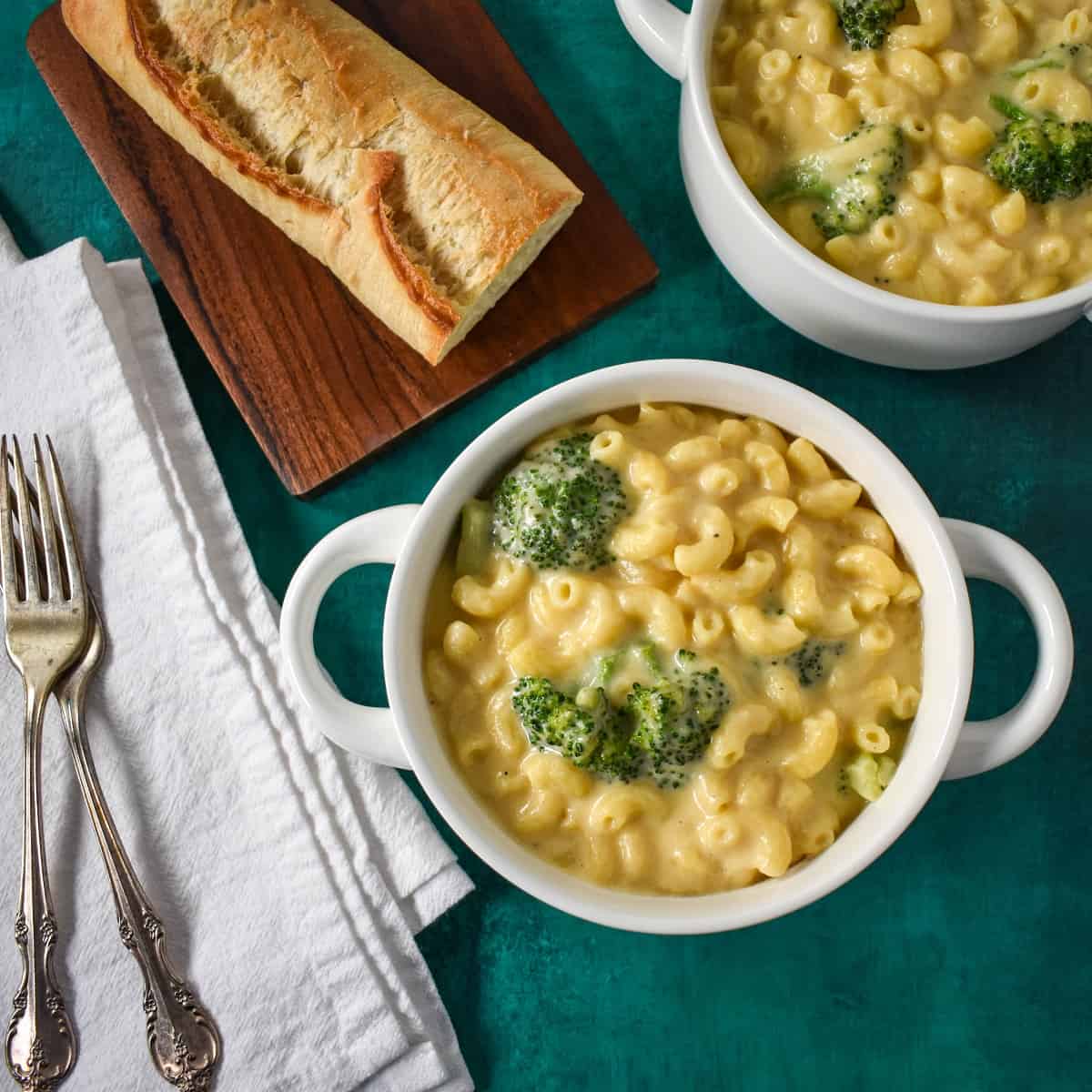 Two white bowls with the broccoli macaroni and cheese set on a green table with a piece of bread and white napkins and forks to the left side.