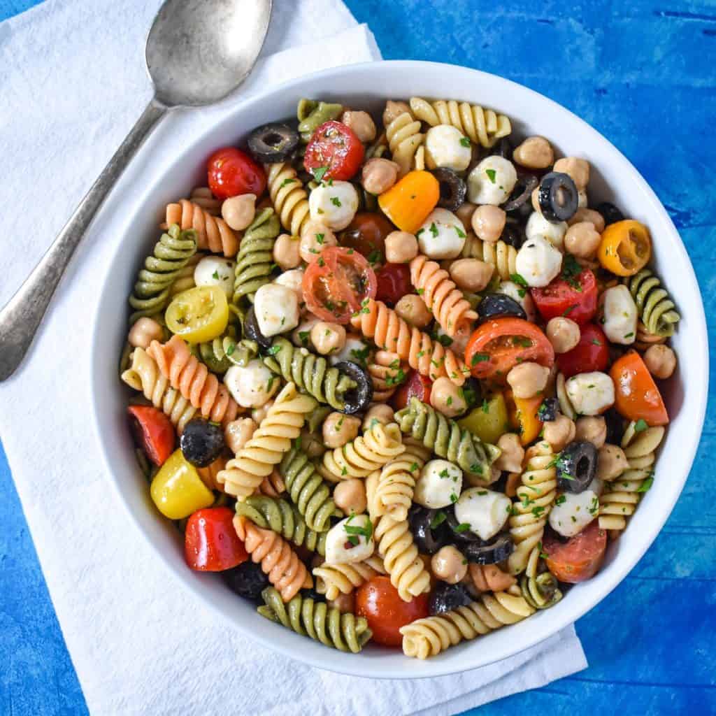 The chickpea pasta salad served in a large white bowl and set on a blue table with a white linen and serving spoon to the left.