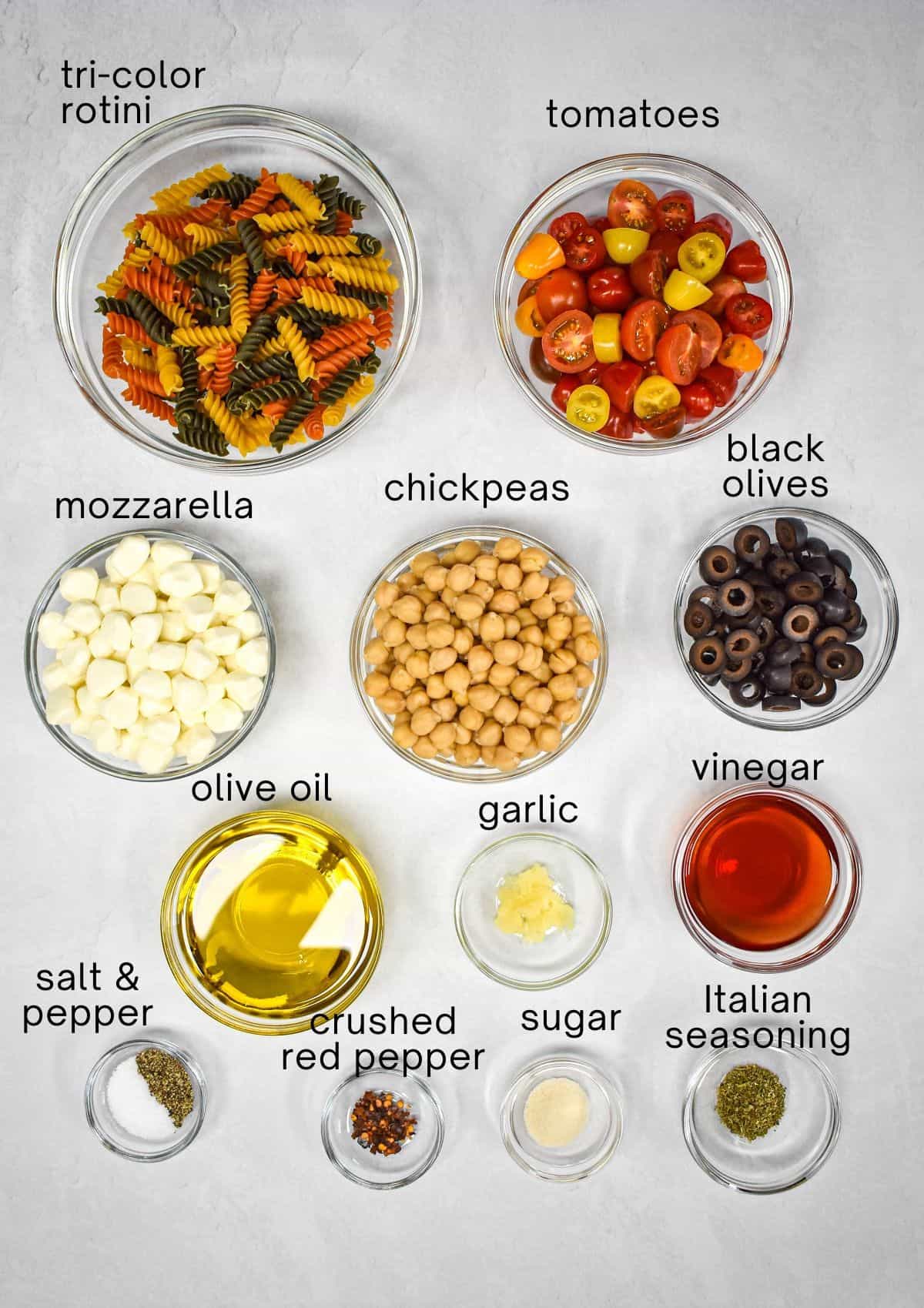 The ingredients for the salad prepped and arranged in glass bowls on a white table with each labeled with small, black letters.
