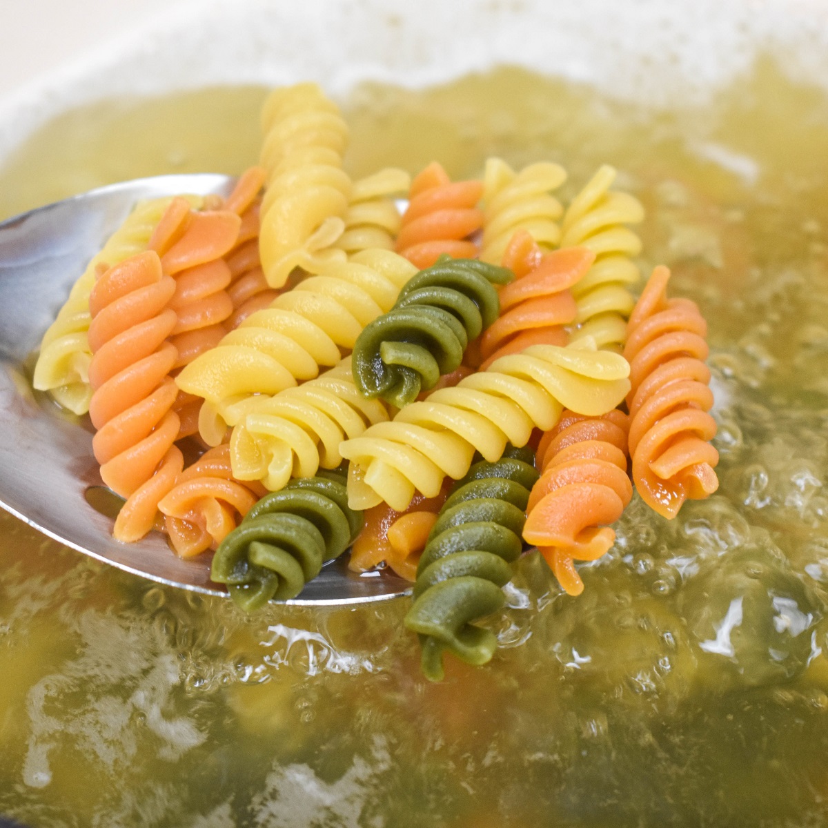Tri-color rotini pasta on a large slotted spoon over a pot of boiling water.
