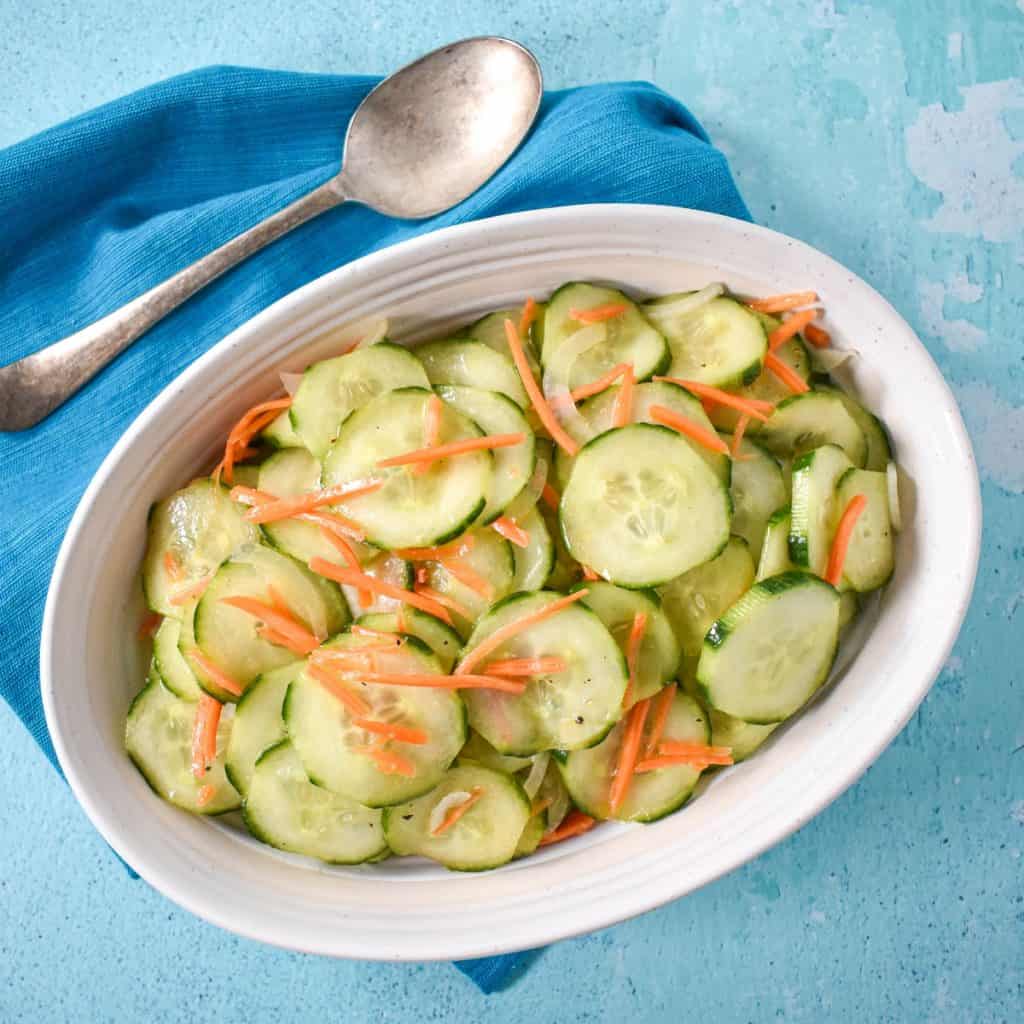 The cucumber salad served in a white bowl and set on a light blue table with a teal linen and serving spoon.