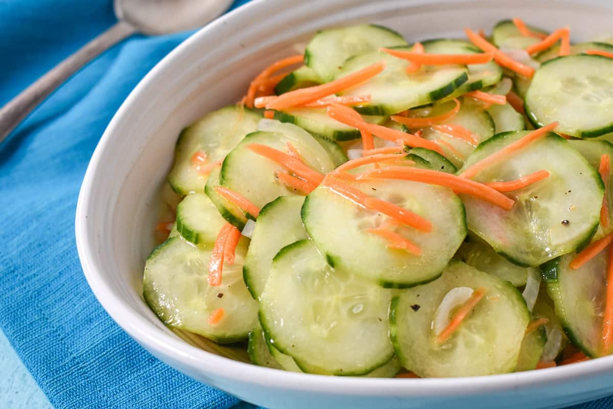 The cucumber salad served in a white bowl and set on a light blue table with a teal linen and serving spoon.