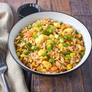 Egg fried rice served in a white bowl set on a wood table with a beige linen and fork to the left.