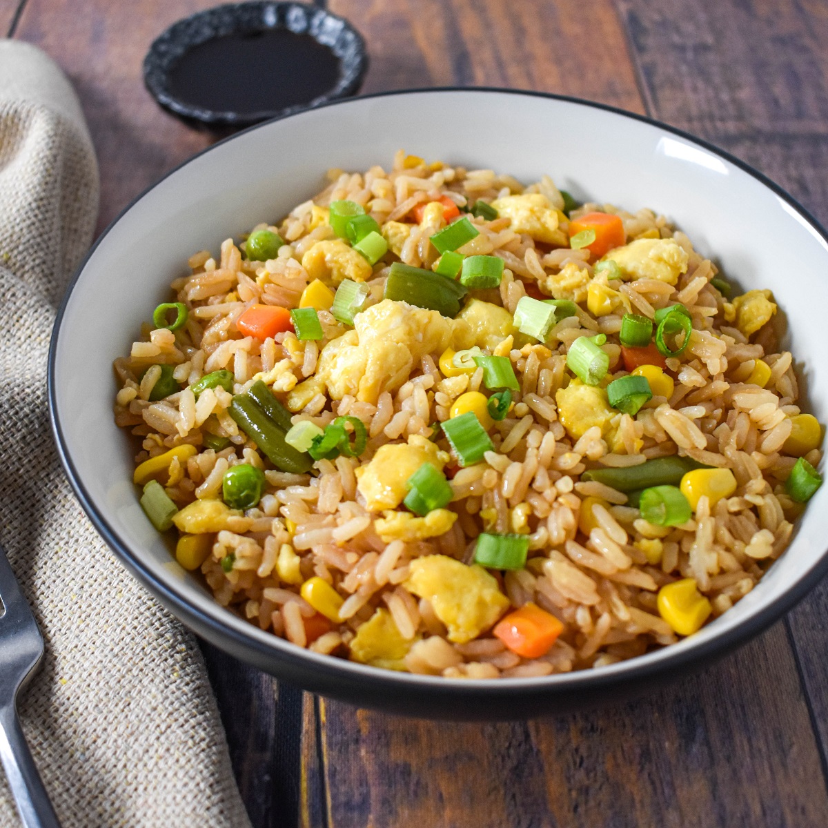 Egg fried rice served in a white bowl set on a wood table with a beige linen and fork to the left.