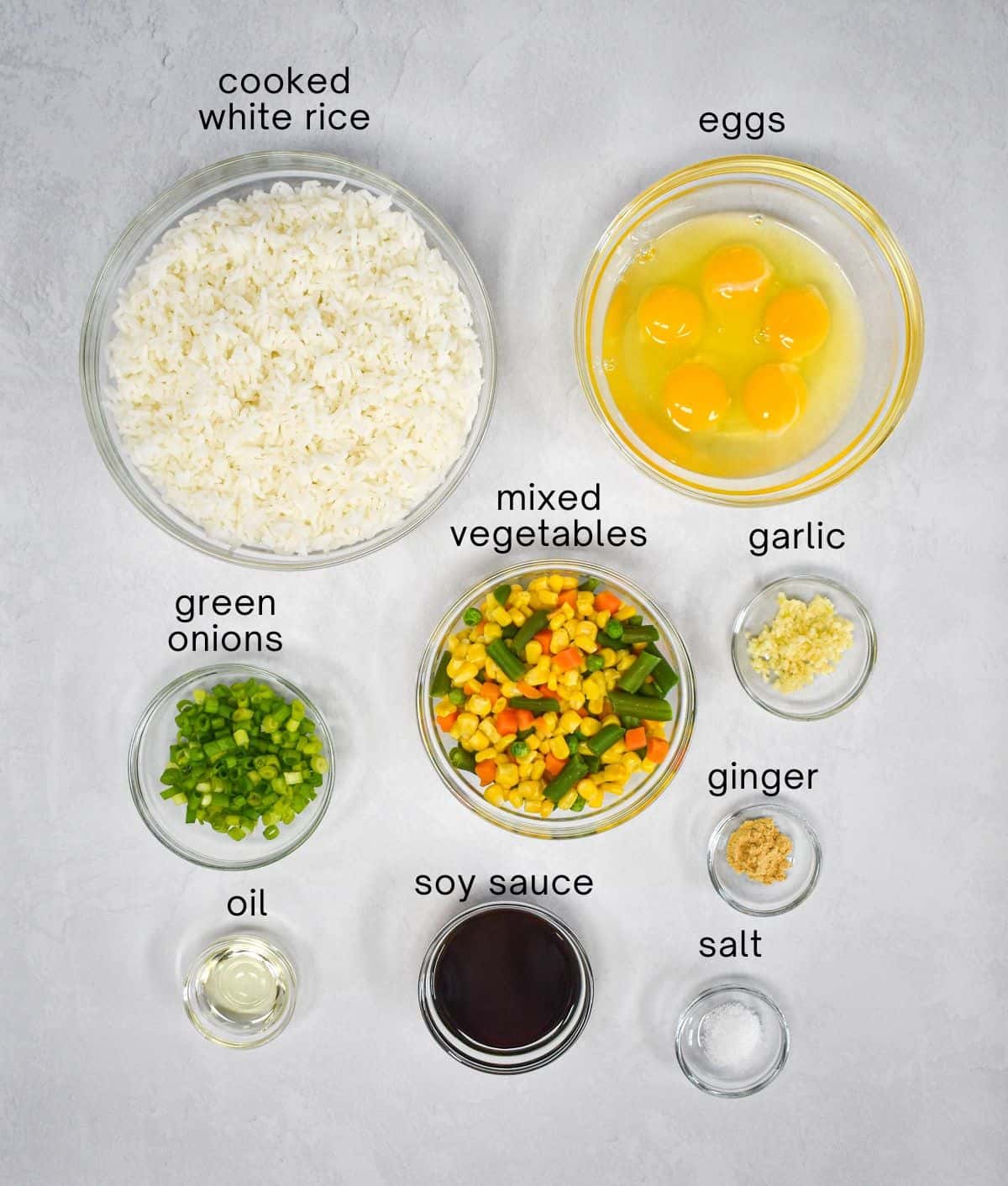 The ingredients for the fried rice arranged in glass bowls on a white table with each labeled with small black letters.