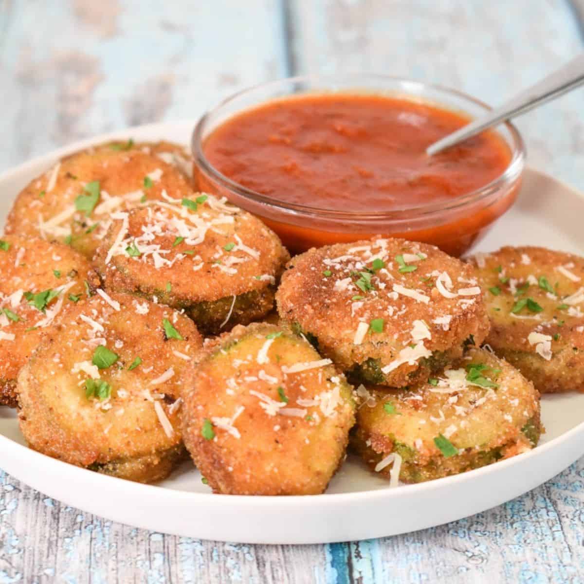 Fried zucchini rounds arranged on a white plate with a small bowl of marinara sauce.