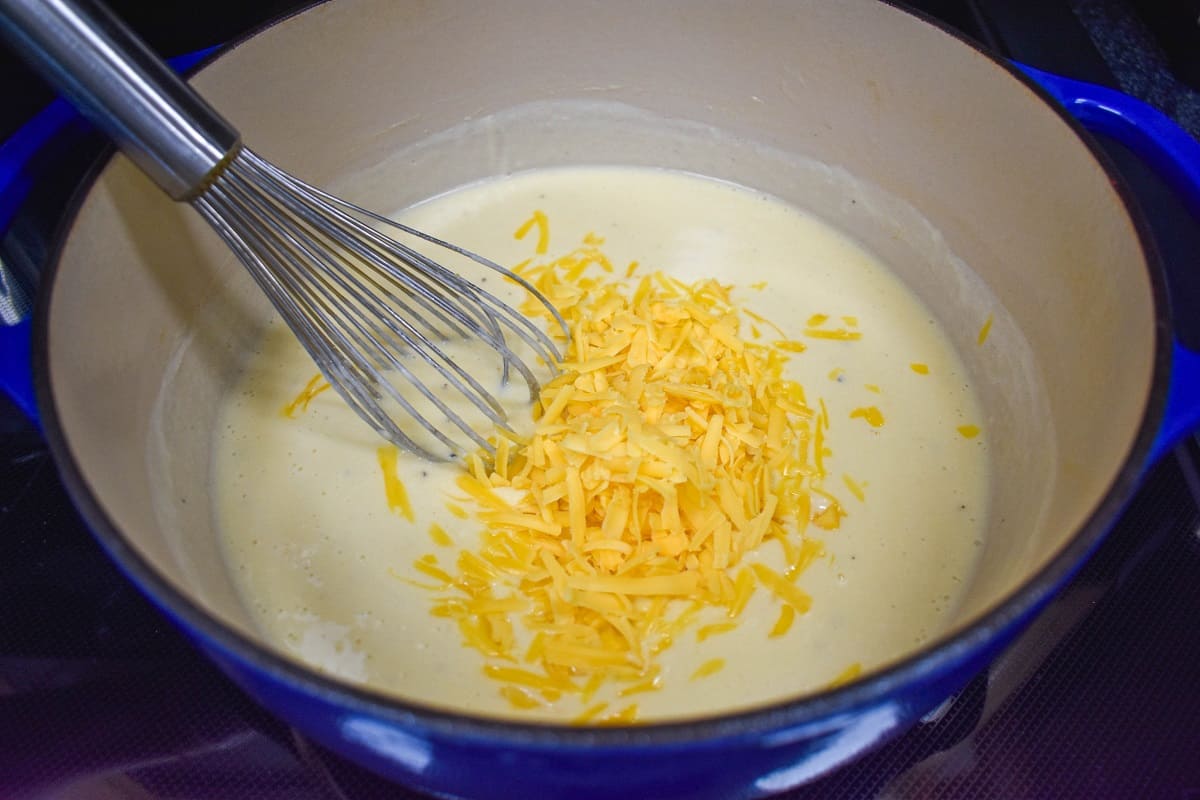 Shredded cheddar cheese being added to the cream sauce in a large pot with a whisk to the left side.