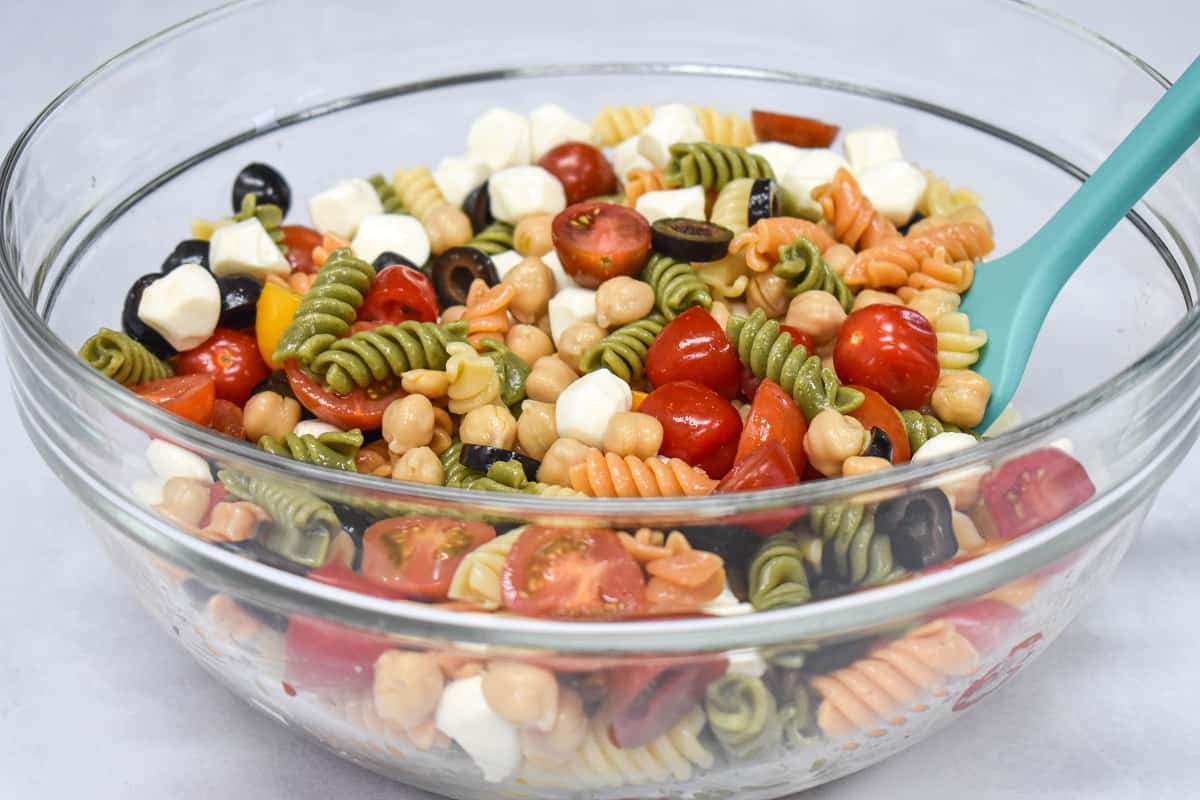 The ingredients for the chickpea pasta salad combined in a large, glass bowls with a teal spatula.