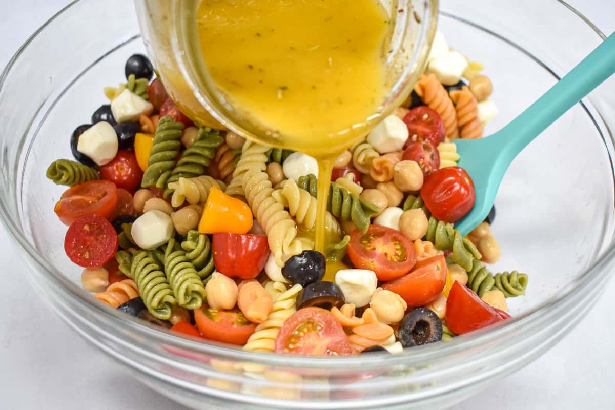 Dressing being added to the ingredients in a large, glass bowl with a teal spatula in the bowl to the right.