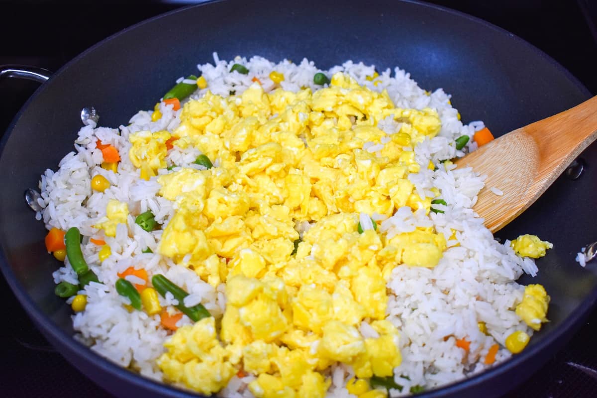 Scrambled eggs on top of the white rice and vegetables in a large skillet with a wooden spoon to the right.