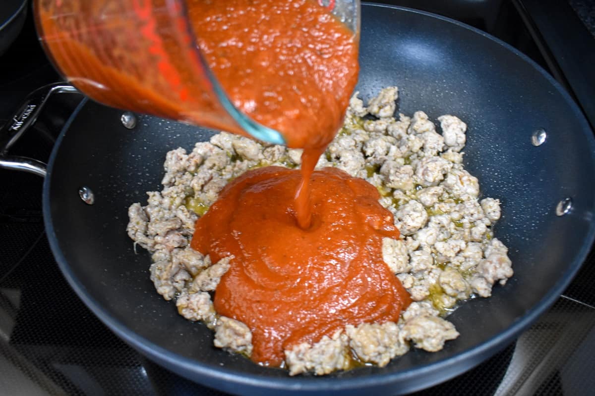 Pasta sauce being added to the browned sausage in a large, black skillet.