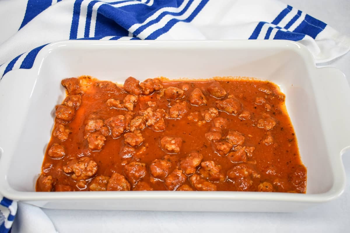 A layer of sausage pasta sauce coating the bottom of a white baking dish and a blue and white kitchen towel in the background. 