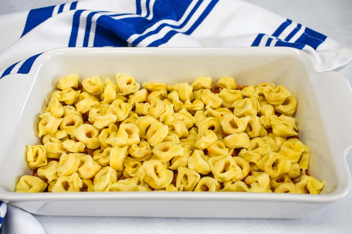 A layer of cooked tortellini in a white baking dish with a white and blue kitchen towel in the background.