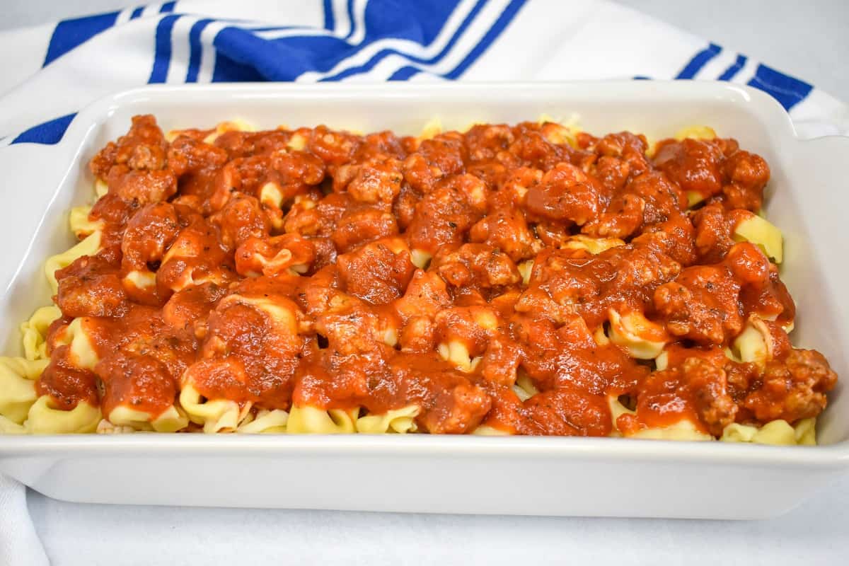 Layers of tortellini and Italian sausage pasta sauce in a white baking dish with a blue and white kitchen towel in the background.