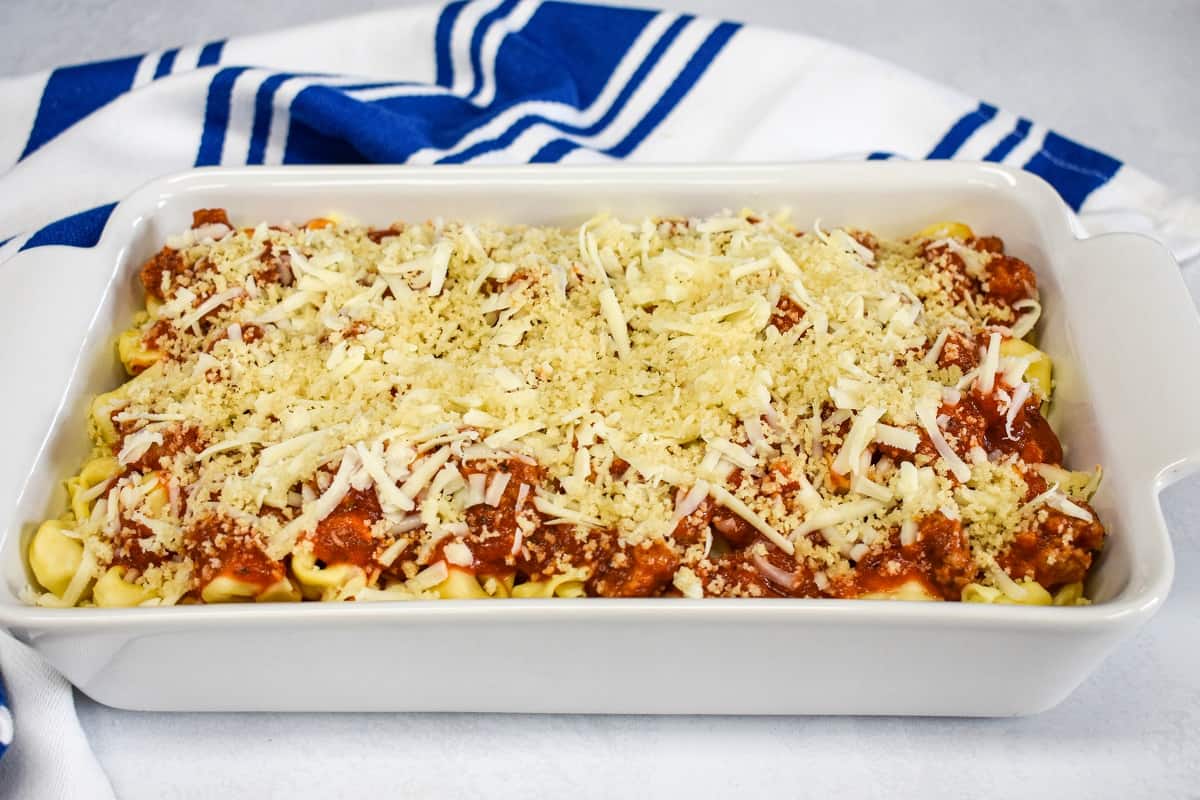 The built tortellini casserole, before baking, in a white dish with a blue and white kitchen towel in the background.