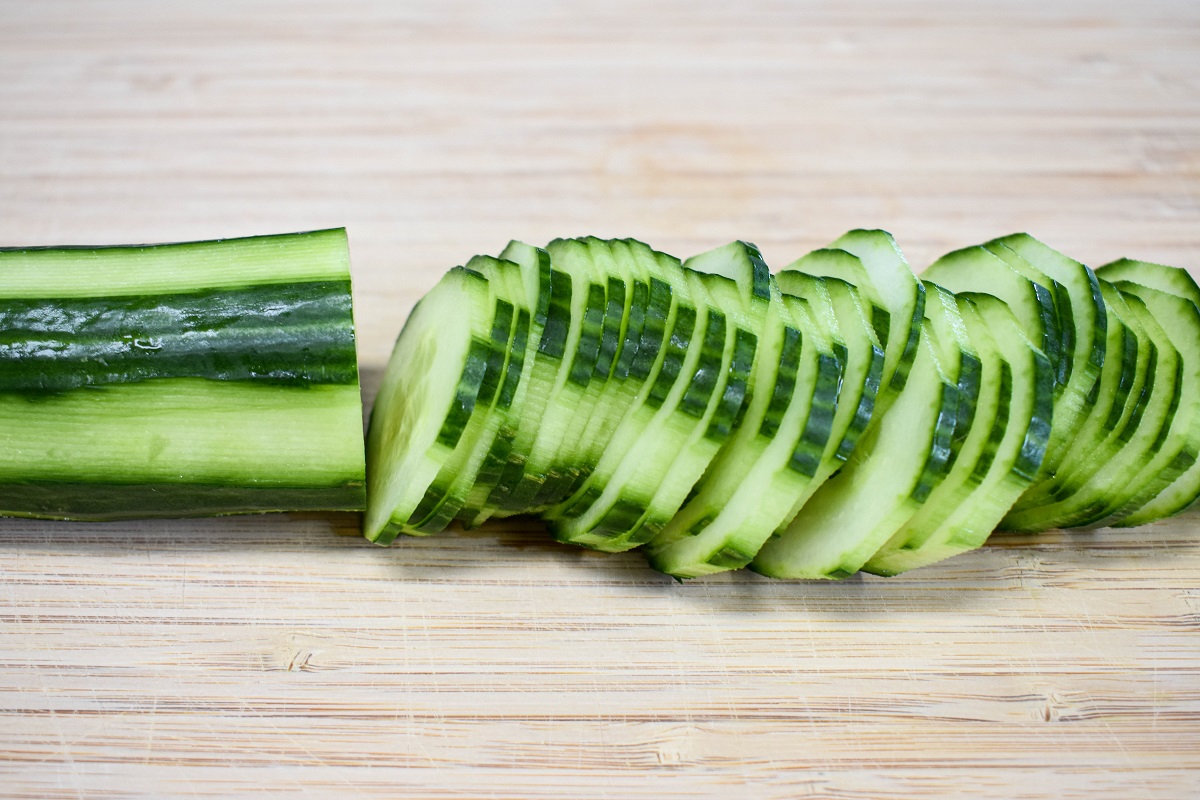 A half peeled and half sliced English cucumber on a wood cutting board.