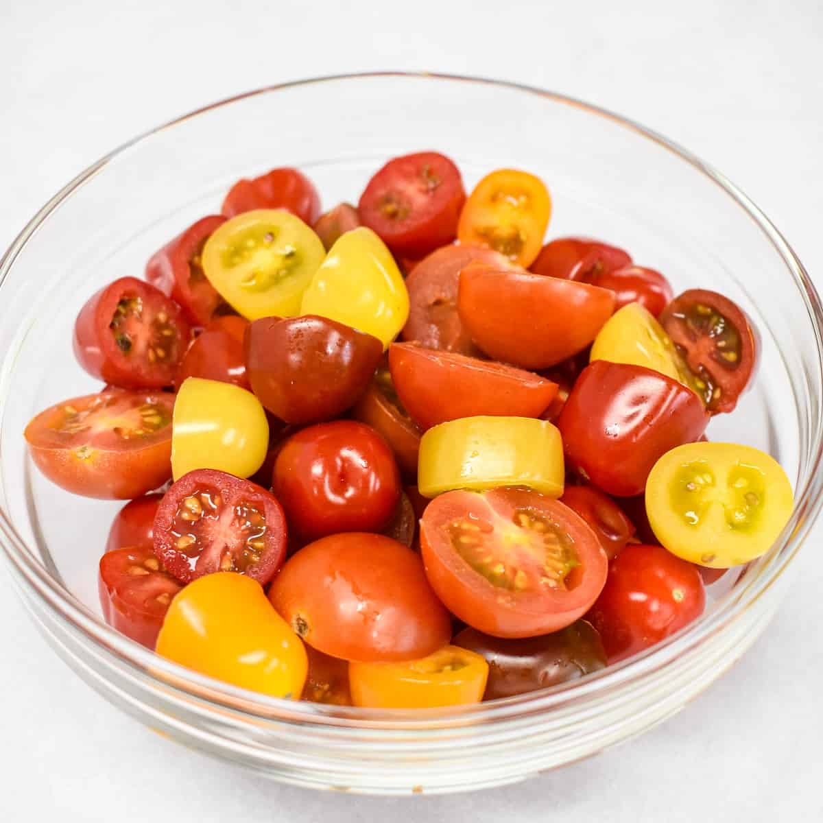 Red and yellow small tomatoes cut in half in a glass bowl on a white table.