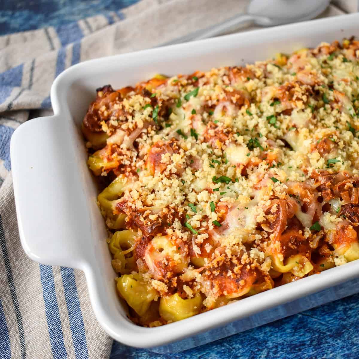 The finished tortellini bake, garnished with parsley, in a white casserole dish set on a blue table with a beige and blue linen.