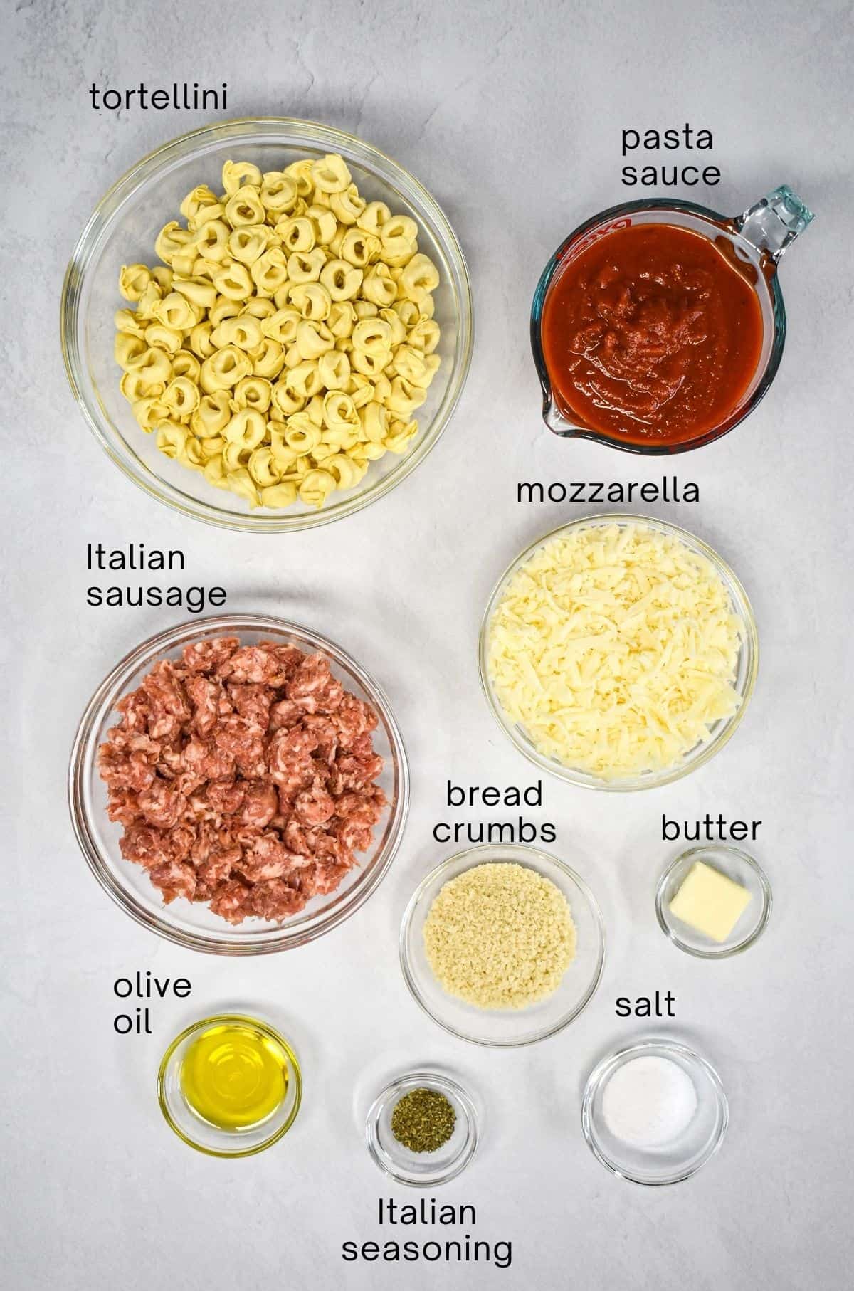 The ingredients for the dish arranged in glass bowls, set on a white table, with each labeled in small, black letters.