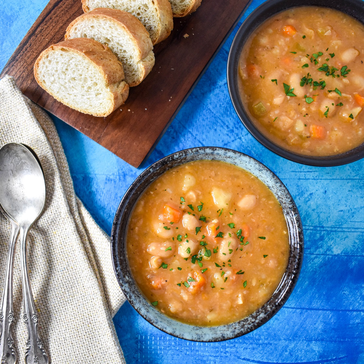 The white bean soup served in a blue bowl, set on a blue table, with a beige linen to the left, and bread and another soup bowl partially showing in the background.