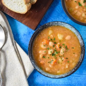 The white bean soup served in a blue bowl, set on a blue table, with a beige linen to the left, and bread and another soup bowl partially showing.