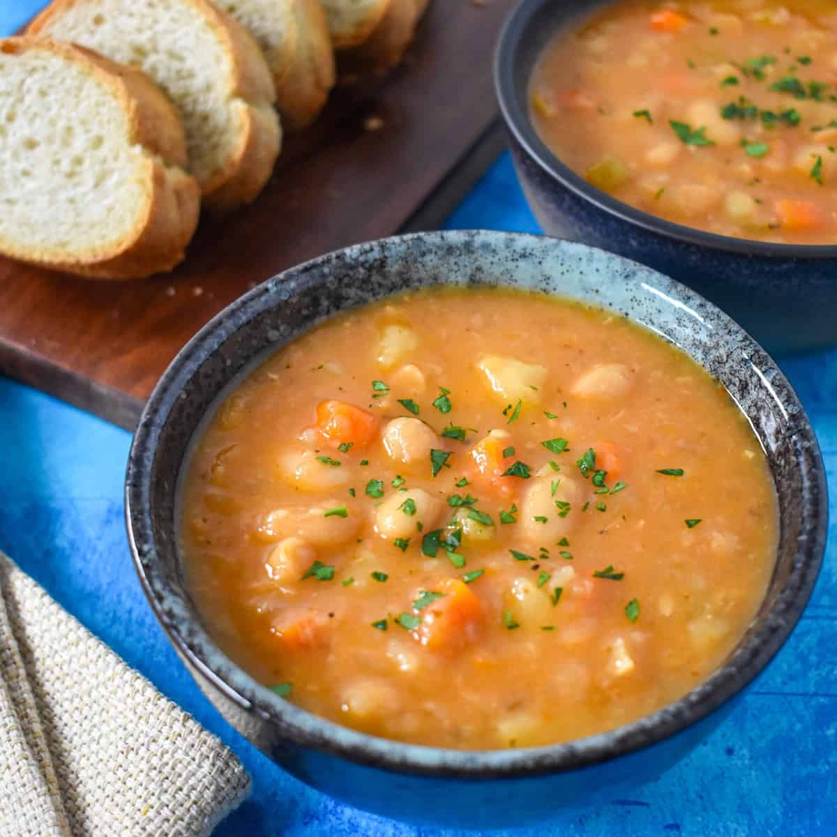 A close-up image of the finished white bean soup served in a blue bowl, garnished with chopped parsley with sliced bread and another bowl of soup partially visible in the background.