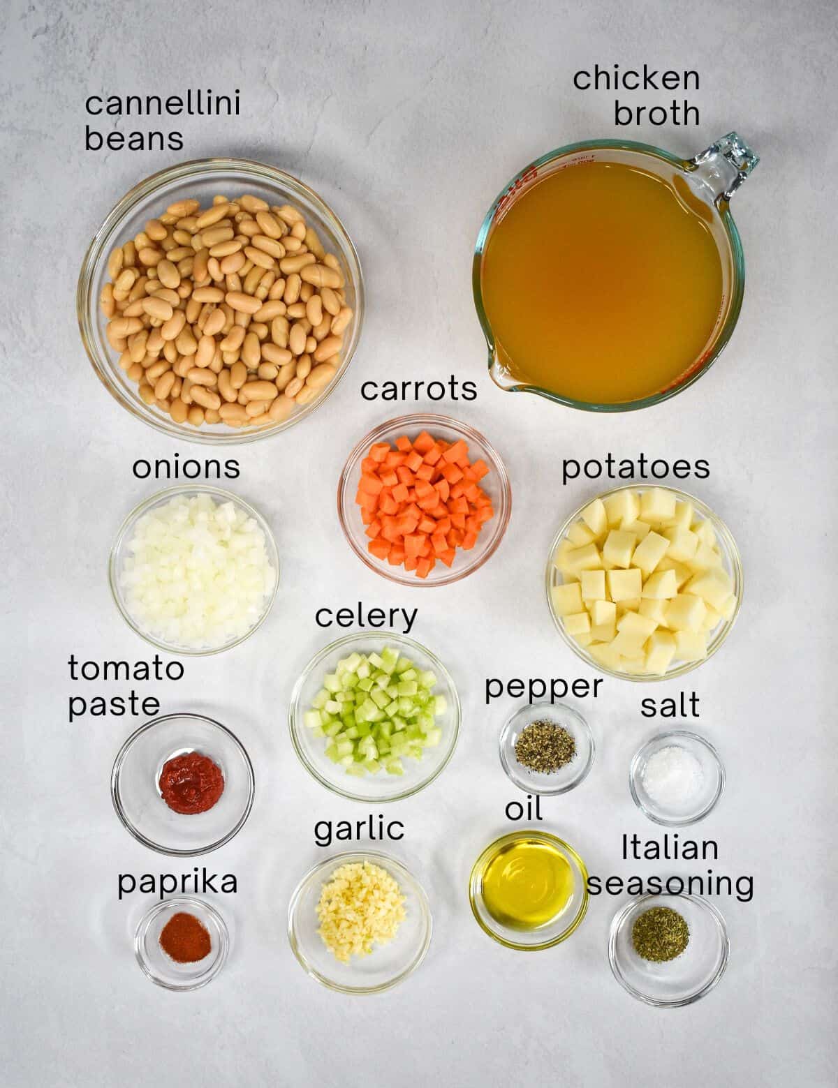 The ingredients for the white bean soup prepped and arranged in glass bowls on a white table with each labeled with small, black letters.